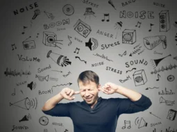 a man standing with his fingers in his ears to stop the influx of information . The source of the information is written on a white wall behind him. It illustrates the noise problem when trying to focus.